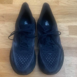 Svarta Hoka Clifton 9 sneakers - Säljer ett par svarta Hoka Clifton 9 sneakers med snyggt mönstrad mesh-ovandel och tjock, dämpad sula. Skorna har snörning och är perfekta för dig som gillar att springa eller promenera långt. Riktigt bekväma och lätta på foten.passar mig och brukar ha 42,5 i skor! Nypris 1400-1500 och knappt använda