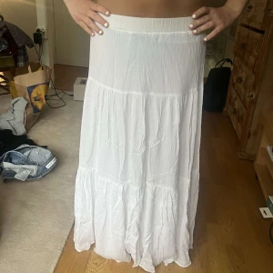 Vit långkjol i bomull - Superfin vit långkjol med volangdetaljer och flowig känsla. Kjolen har en enkel midja och är tillverkad i ett lätt, krinklat bomullstyg som ger en avslappnad och luftig look. Perfekt för varma dagar och enkel att styla med crop tops eller stickade tröjor.
