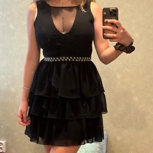 Svart volangklänning med strassdetalj - Supersnygg svart kortklänning med flera volanger i kjolen och markerad midja med strassbälte. Djup v-ringning med meshdetalj upptill och ärmlös design. Perfekt för dig som vill sticka ut med en elegant och trendig look.