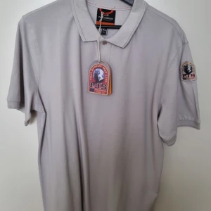 Stone blue pikétröja från Parajumpers gangapuma - Snygg ljusgrå pikétröja från Parajumpers med klassisk krage, korta ärmar och broderad patch på ärmen. Tillverkad i bomull med diskreta slitsar i sidorna och orange detaljsöm. Perfekt för en clean och stilren look.