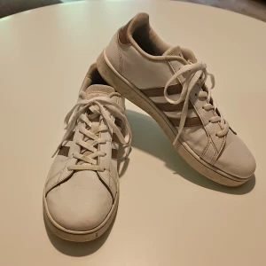 Vita Adidas sneakers med bruna ränder - Snygga vita Adidas sneakers med klassiska tre bruna ränder på sidorna. Skorna har snörning, rund tå och platt sula med hexagonmönster undertill. Tillverkade i syntetmaterial och textil för en clean och sportig look.