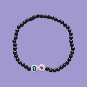 Svart pärlarmband med bokstav och hjärta - Stilrent svart pärlarmband med små runda pärlor och två vita pärlor framtill, en med bokstaven D och en med ett rött hjärta. Perfekt accessoar för att ge din outfit en personlig touch.