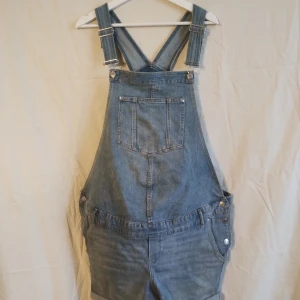 Jeansshorts hängselshorts MAMA XL - Blå jeansshorts med hängslen från MAMA i storlek XL. Klassisk denimlook med stora fickor fram och bak, justerbara axelband och upprullade benslut. Perfekt för dig som gillar en avslappnad och cool stil.
