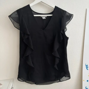 Svart blus med volang och v-ringning - Snygg svart blus med v-ringad hals och volangdetaljer längs sidorna. Ärmlös modell med transparenta partier vid axlarna som ger en luftig känsla. 