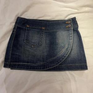 Mörkblå jeanskjol med ficka - Säljer denna sygga mörkblå jeanskjol i kort modell med cool asymmetrisk söm framtill och två knappar i midjan. Klassisk ficka fram och snygga detaljer bak. Inga tecken på användning.