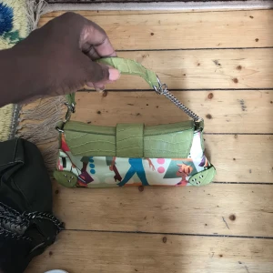 Grön axelväska med färgglatt mönster - Säljer en unik axelväska med gröna detaljer i croco-imitation och kedjehandtag. Väskan har ett färgglatt, abstrakt mönster i blått, rött, rosa och orange. Snygga metalldetaljer och cool form som sticker ut. Perfekt för dig som vill ha något annorlunda!