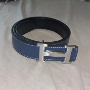 H blått skärp med H-spänne - H bälte som man kan vända håll och göra till svart om man vill.Snyggt blått skärp från H med ikoniskt H-spänne i silverfärgad metall. Bältet är gjort i slätt skinn med vita kontrastsömmar och har en stilren, klassisk design som sticker ut. Perfekt accessoar för att lyfta din outfit.