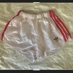 Vita Adidas shorts med röda ränder - Snygga vintage Adidas shorts i vitt med klassiska röda ränder längs sidorna och elastisk midja. Lätta och glansiga i materialet, perfekta för träning eller chill. Adidas-logga framtill och bakficka för extra stilpoäng.