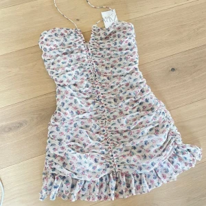 Blommig kortklänning med volang - Supersöt kortklänning från ZARA med blommigt mönster i rosa och blått på vit botten. Klänningen har smala axelband, rynkade detaljer framtill och volangkant nertill. Perfekt för sommarens alla tillfällen!