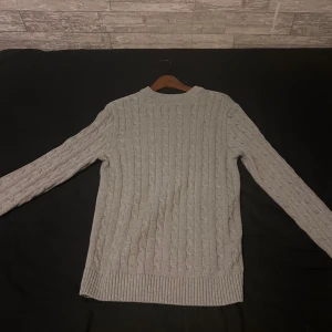 Grå kabelstickad tröja - Snygg grå kabelstickad tröja med rund hals och långa ärmar. Tröjan har klassiskt flätmönster och breda ribbade muddar vid ärmslut och nederkant. Perfekt för lager-på-lager och chill höst- eller vinterstil.