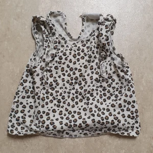 Topp - Supersöt ärmlös topp från H&M i storlek 68 med leopardmönster i svart och brunt på beige botten. Toppen har volangdetaljer vid axlarna och knäppning bak i nacken för enkel påklädning. Perfekt för dig som gillar djurmönster och vill ha något extra gulligt.
