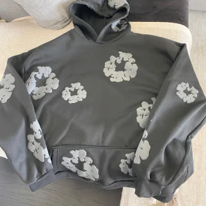 Svart hoodie från denim tears - Cool svart hoodie med stora gråa blommönster över hela tröjan. Har huva och magficka, samt ribbade muddar vid ärmslut och nederkant. Perfekt för dig som gillar streetstyle och vill sticka ut lite extra.