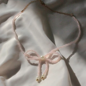 Pärlhalsband med rosett  - Supergulligt halsband med små ljusrosa pärlor formade till en rosett, dekorerat med vita pärlor och guldfärgade detaljer. Perfekt för dig som gillar söta accessoarer med en feminin touch.