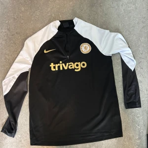 Chelsea Nike långärmad träningsset - Chelsea FC träningsset från Nike med långärmad svart tröja och svarta byxor. Tröjan har vita partier på axlar och ärmar, guldiga loggor och 'trivago' tryckt på bröstet. Byxorna har Chelsea-märke och Nike-logga i guld. Materialet är lätt och andas, perfekt för träning.