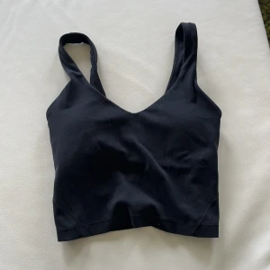 Svart linnetopp från Lululemon - Säljer en svart linnetopp från Lululemon med breda axelband och v-ringad front. Toppen är croppad och har en stilren, sportig look med diskret logga framtill. Perfekt för träning eller chill dagar. Mjukt och stretchigt material som sitter skönt mot kroppen.