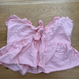 Rosa bandeau topp med knyt - Supersöt ljusrosa bandeau topp med volangkant och knyt framtill. Toppen är ärmlös och har smockad rygg för extra komfort. Perfekt för varma dagar och ger en romantisk vibe till din outfit.