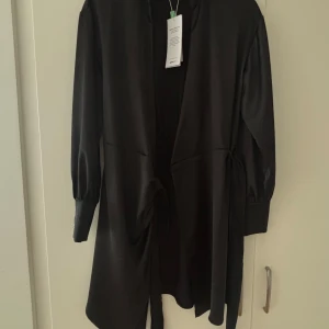 Svart omlottklänning i satin XS - Supersnygg svart omlottklänning i glansig satin från GinaTricot Good Collection. Klänningen har långa ärmar, krage och knytband i midjan för en stilren look. Perfekt för dig som gillar minimalistisk och elegant stil.