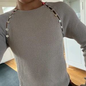 Ribbad grå tröja med knappar från Zara - Supersnygg ribbad grå tröja från Zara med lång ärm och coola knappdetaljer längs axlarna. Tröjan har en tight passform och är tillverkad i mjukt stickat material. Perfekt för dig som gillar unika detaljer och stilren look.🍒❣️🍏