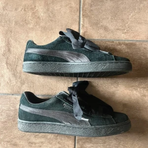 Svarta Puma Suede sneakers med breda band - Snygga svarta Puma Suede sneakers med breda svarta band som snörning. Skorna har klassisk låg profil, rund tå och är gjorda i mjuk mocka med svarta detaljer och sula. Perfekta för dig som gillar streetstyle och vill ha en ikonisk look. Storlek 38,5. 