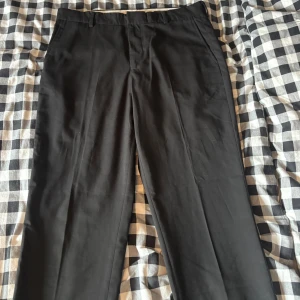 Svarta kostymbyxor från H&M Regular Fit - Klassiska svarta kostymbyxor från H&M i modellen Regular Fit. Byxorna har raka ben, bälteshällor och diskreta bakfickor. Materialet är en mix av polyester och viskos med lite elastan för extra komfort. Perfekta för en clean och stilren look.