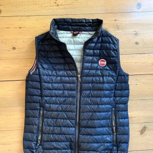 Colmar väst  - Snygg blå  dunväst från Colmar med quiltad design och hög krage. Västen har dragkedja framtill, två fickor med dragkedja och ett broderat Colmar-märke på bröstet. Insidan är ljusgrå och den har en sportig känsla med diskreta ränder vid ärmöppningarna.