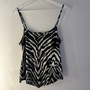 Zebramönstrad linnetopp Gina Tricot - Säljer en snygg zebramönstrad linnetopp från Gina Tricot i svart och vitt. Toppen har tunna axelband och är i ett lätt, mjukt material som känns skönt mot huden. Perfekt för dig som vill sticka ut med ett djurmönster och gillar trendiga detaljer. Två små hål vid bandet, se bild 4 o 5