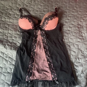 Svart och rosa spetsklänning - Söt kort klänning i svart mesh med rosa detaljer och vadderade kupor. Klänningen har spets längs kuporna och framtill, samt små prickar i tyget. Axelbanden är justerbara och nederkanten har volang. Perfekt för dig som gillar romantisk och feminin stil. Lite böjd i ena cupan men syns inte när man har den på sig 