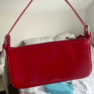 Röd axelväska från Gina Tricot - Röd axelväska i glansigt syntetmaterial. Använt ett fåtal gånger