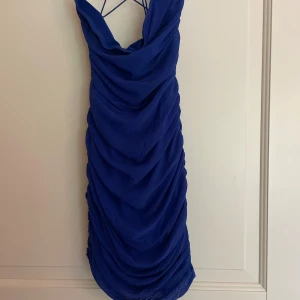 Blå draperad meshklänning med snörning - Snygg blå kortklänning i mesh med draperad design och tunna axelband med snörning i ryggen. Klänningen har en tight passform och är perfekt för dig som vill sticka ut. Materialet är lätt och stretchigt, vilket gör den bekväm att bära hela kvällen.