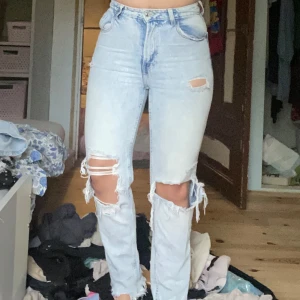 Ljusblå ripped highwaist jeans - Säljer ett par ljusblå jeans med hög midja och raka ben. Jeansen har flera slitningar och hål vid knäna och låren för en riktigt trendig, avslappnad look. Klassisk femficksmodell och stängs med dragkedja och knapp.