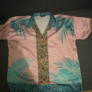 Rosa skjorta med leopard- och palmtryck - Färgstark kortärmad skjorta från Man i 100% polyester. Skjortan är rosa med turkosa palmblad, leopardmönster längs knäppningen och kragen samt detaljer i blått leopardmönster. Perfekt för dig som gillar unika och vågade plagg.