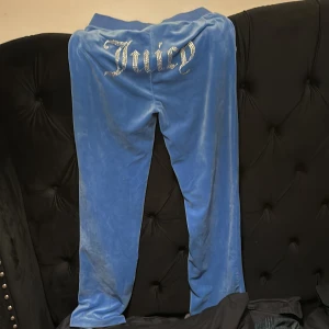 Blå velourbyxor Juicy Couture S - Blåa mjukisbyxor från Juicy Couture i mjuk velour med snörning i midjan och glittrande 'Juicy' text bak. Klassisk relaxed fit och ikonisk logga framtill. Perfekta för en chill och trendig look.