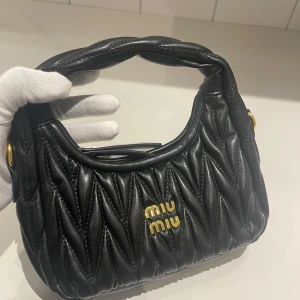 Svart quiltad handväska från Miu Miu - Säljer en svart quiltad handväska från Miu Miu i skinn med guldiga detaljer och logga framtill. Väskan har en mjuk, vadderad struktur och ett kort handtag. Stängs med dragkedja och har en lyxig vibe som sticker ut.