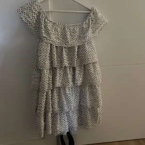 Prickig volangklänning från Zara - Supersöt kort offshoulder-klänning från Zara i vitt med svarta prickar. Klänningen har flera volanger, en svart knytbälte i midjan och är tillverkad i ett lätt, luftigt material. Perfekt för sommarens alla tillfällen!
