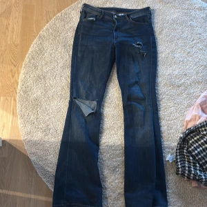 Mörkblå bootcut jeans med slitningar - Säljer ett par mörkblå jeans med bootcut passform och snygga slitningar på låret och knät. Jeansen har klassisk femficksdesign och stängs med dragkedja och knapp. Perfekta för en avslappnad och trendig look.