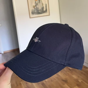 Mörkblå keps från GANT  - Snygg mörkblå keps från GANT med klassisk böjd skärm och broderad logga framtill. Kepsen är tillverkad i ett mjukt och slitstarkt bomullstyg och har en stilren look som passar till det mesta. Passar alla huvudformer/storlekar eftersom den går att justera där bak. Säljer den för 200kr, nypris ligger på 550kr. 