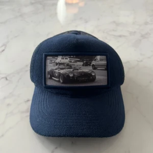 Blå keps från Emilio Pendenza med bilmotiv - Snygg blå truckerkeps från Emilio Pendenza med mesh baktill och justerbar snapback. Framtill finns en stor patch med svartvit bild på en klassisk sportbil. Perfekt accessoar för dig som gillar bilar och streetstyle. Spännet är sönder men går fortfarande att använda