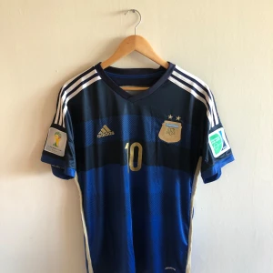 Argentina Messi VM 2014 bortatröja Adidas - Argentina bortatröja från VM 2014 med Messi 10 på ryggen. Djupblå och marinblå med guldiga detaljer, vita Adidas-ränder på axlarna och officiella FIFA World Cup och Football for Hope-patchar på ärmarna. Tillverkad i lätt funktionsmaterial. Finns I storlek M och L.         Tveka inte om du hat mågon fundering!!