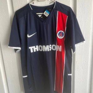 PSG Ronaldinho 10 Nike fotbollströja - Paris Saint-Germain fotbollströja med Ronaldinho och nummer 10 på ryggen. Tröjan är marinblå med en röd vertikal rand och vita detaljer, samt PSG-emblem och THOMSON-tryck på bröstet. Klassisk V-ringning och korta ärmar. Perfekt för dig som älskar retro och fotbollshistoria.