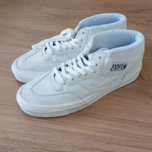Vita Vans Half Cab sneakers mocka storlek 43 - Säljer ett par klassiska Vans Half Cab sneakers i helvitt mocka. Skorna har mellanhögt skaft, vit sula och snörning. Ikonisk Half Cab-logga på sidan och diskret Vans-märke på tungan. Perfekta för dig som gillar clean och stilren streetwear.