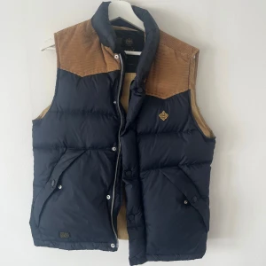 Mörkblå dunväst med manchester - Snygg mörkblå dunväst från Morris med bruna detaljer i manchester på axlarna. Västen har dragkedja och tryckknappar framtill, två stora fickor och ett ljust innerfoder. Klassisk badge på bröstet och quiltad look. Perfekt för lager-på-lager-stil.