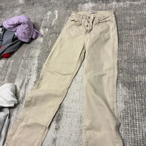 Beiga raka jeans från NA-KD, strl 34 - Snygga beiga jeans från NA-KD i rak modell med hög midja. Byxorna har klassiska fem fickor, knäppning med flera silverfärgade knappar och är tillverkade i bomull. Perfekta till en chill vardagslook.