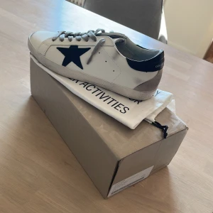 Golden Goose  - Golden Goose sneakers i vitt skinn med svart stjärndetalj på sidan, grå mockadetalj vid hälen och svart hälkappa med logga. Klassisk rund tå, grå snörning och platt sula. Snygg och stilren modell som passar till allt. Hör av er för frågor.