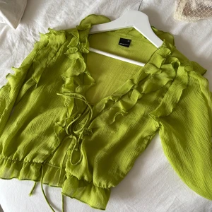 Grön volangblus från Gina Tricot XS - Supersöt ljusgrön blus från Gina Tricot med volanger längs v-ringningen och ärmarna. Blusen har långa, lätt utsvängda ärmar och knytning framtill. Tunt, luftigt material som ger en flowy look💕