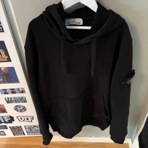 Svart hoodie från Stone Island - Svart hoodie från Stone Island med klassisk logga på ärmen. Tröjan har huva med snörning och en stor magficka framtill. Perfekt för dig som gillar streetwear och vill ha en stilren look. Materialet känns mjukt och skönt, troligen bomull.