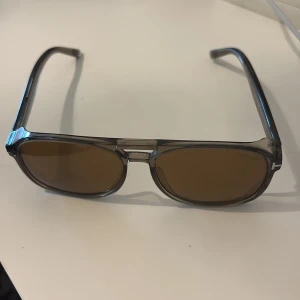 Tom Ford solglasögon i grå plast - Snygga Tom Ford solglasögon med grå transparent båge och bruna linser. Klassisk fyrkantig form med diskret T-detalj vid gångjärnet. Ramen är i plast och ger en modern vibe, perfekt för dig som vill ha en stilren accessoar.