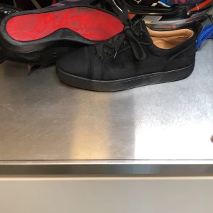 Svarta sneakers från Louboutin - Snygga svarta sneakers från Christian Louboutin i mocka med klassisk röd sula och svarta snören. Skorna har en låg profil och rund tå, samt en stilren design som passar till många outfits. Insidan är beige och sulan har den ikoniska Louboutin-signaturen.