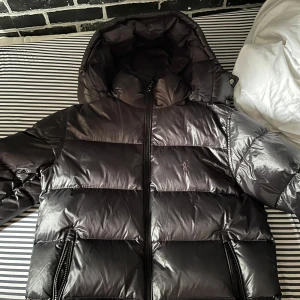 Svart pufferjacka från Polo Ralph Lauren - Säljer en svart pufferjacka från Polo Ralph Lauren med glansig finish och broderad logga på bröstet. Jackan har huva, dragkedja framtill och två fickor med dragkedja. Perfekt för kalla dagar. Köpt på NK för 6000kr nypris och är sparsamt använd. Unik med den svarta loggan vilket man inte ser så ofta. Priset är ej hugget i sten