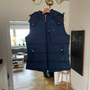 Dunväst - Säljer en marinblå dunväst från Jacson med huva och dragkedja framtill. Västen har två fickor med dragkedja och en diskret Jacson-logga på bröstet. Perfekt för lager-på-lager och passar dig som gillar ridning eller vill ha en sportig look. Västen är i storlek L men skulle säga att den passar dig som är en M. Använd fåtal gånger.