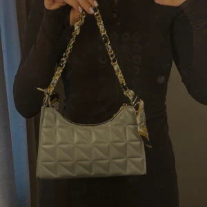 Axelväska i ljusgrått skinn - Säljer en snygg quiltad axelväska i ljusgrått skinn med kedjehandtag flätat med mönstrad scarf. Väskan har dragkedja upptill och geometriskt mönster på framsidan. Perfekt accessoar för att lyfta din outfit.